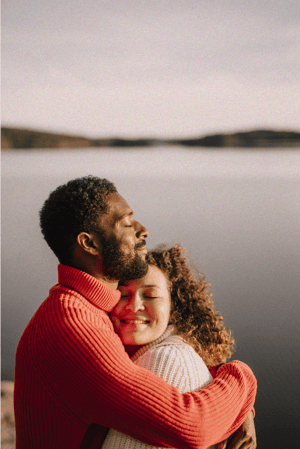 Man and woman lovingly embracing in front of a body of water with gentle smiles on their faces