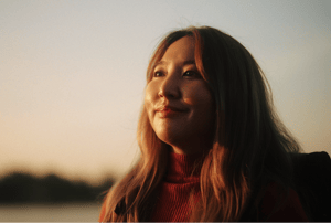 A woman with long hair smiling softly while looking into the distance during sunset, with a calm outdoor background