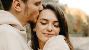 A man kissing a woman on the temple as she smiles softly with her eyes closed, nestled in his arms outdoors.
