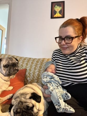Becki on a sofa holding newborn Betsy, with her two pugs sitting beside her