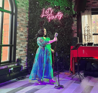 Becki singing into a microphone in a green dress at an event with a "Let's Party" neon sign