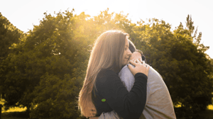 A heterosexual couple embracing in front of some trees and the sun is beginning to set