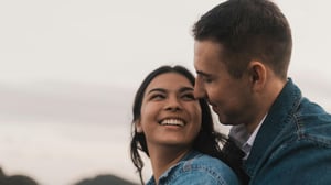 A woman looking up and laughing as a man leans in close, both smiling against an overcast sky