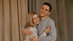 A smiling couple embracing indoors, the man hugging the woman from behind as they both laugh