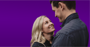A couple smiling at each other with a purple backdrop behind.